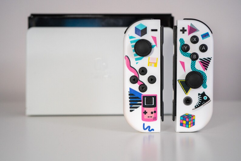 May include: A white Nintendo Switch controller with a retro 80s and 90s themed design. The controller features a variety of colorful geometric shapes, including a pink Game Boy, a blue and white sneaker, and a rainbow Rubik's Cube.
