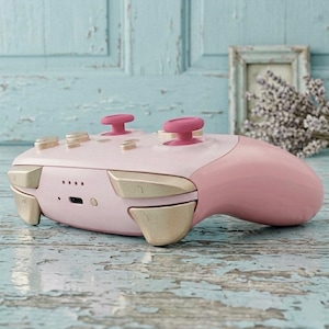 May include: A wireless video game controller featuring a light pink body, two vibrant pink analog sticks, and metallic gold shoulder buttons and triggers. The device includes a central USB-C charging port and four small LED indicator lights.