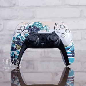 May include: A white and black PlayStation 5 controller with a custom design featuring a blue and white wave pattern. The controller has black joysticks and buttons, with the PlayStation logo centered. The design is inspired by Japanese art.