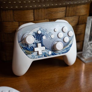 May include: White and blue video game controller featuring a wave design. The controller has a D-pad, buttons, and analog sticks. The wave design is a classic Japanese artwork. The controller is on a wooden surface.