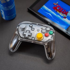 May include: A transparent gaming controller with gray buttons, a yellow center button, and red and green buttons. A blue energy drink and a gaming console with a Zelda game are in the background. The controller is on a dark surface.