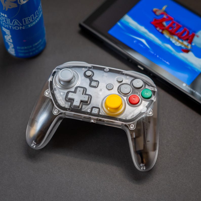 May include: A transparent gaming controller with gray buttons, a yellow center button, and red and green buttons. A blue energy drink and a gaming console with a Zelda game are in the background. The controller is on a dark surface.