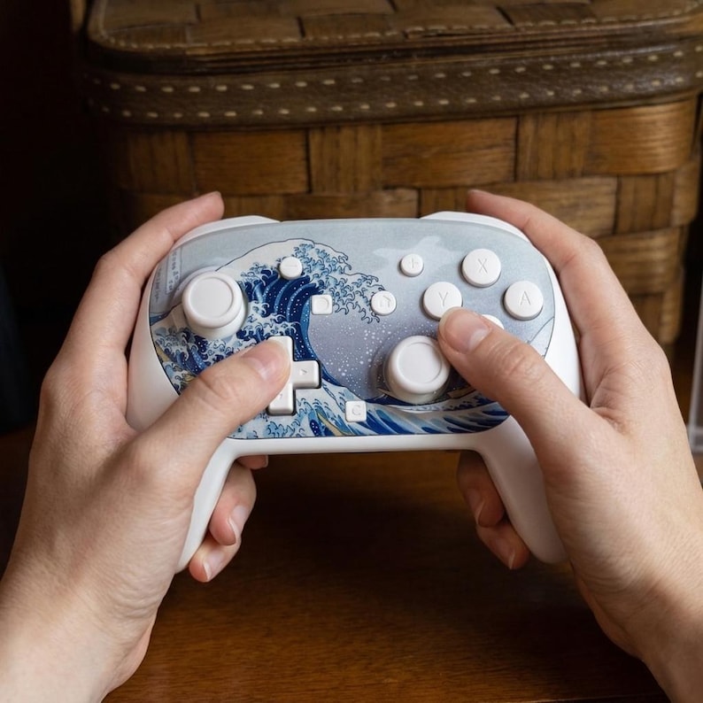 May include: White gaming controller featuring a blue and white wave design. The controller has a D-pad, face buttons, and analog sticks. The design is inspired by the famous Japanese artwork, "The Great Wave off Kanagawa."