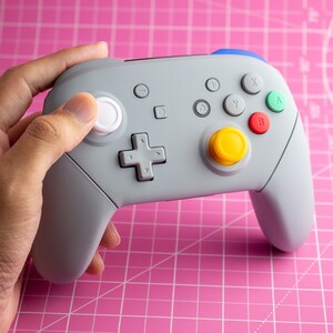 May include: A gray video game controller with a white button, yellow, red, and green buttons, and a directional pad. The controller is on a pink surface.
