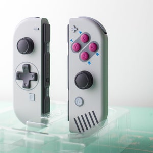 Może przedstawiać: Dwa szaro-białe kontrolery Nintendo Switch z r&oacute;żowymi i niebieskimi przyciskami. Kontrolery mają retro design z czarnym przyciskiem krzyża i czarnym przyciskiem koła.
