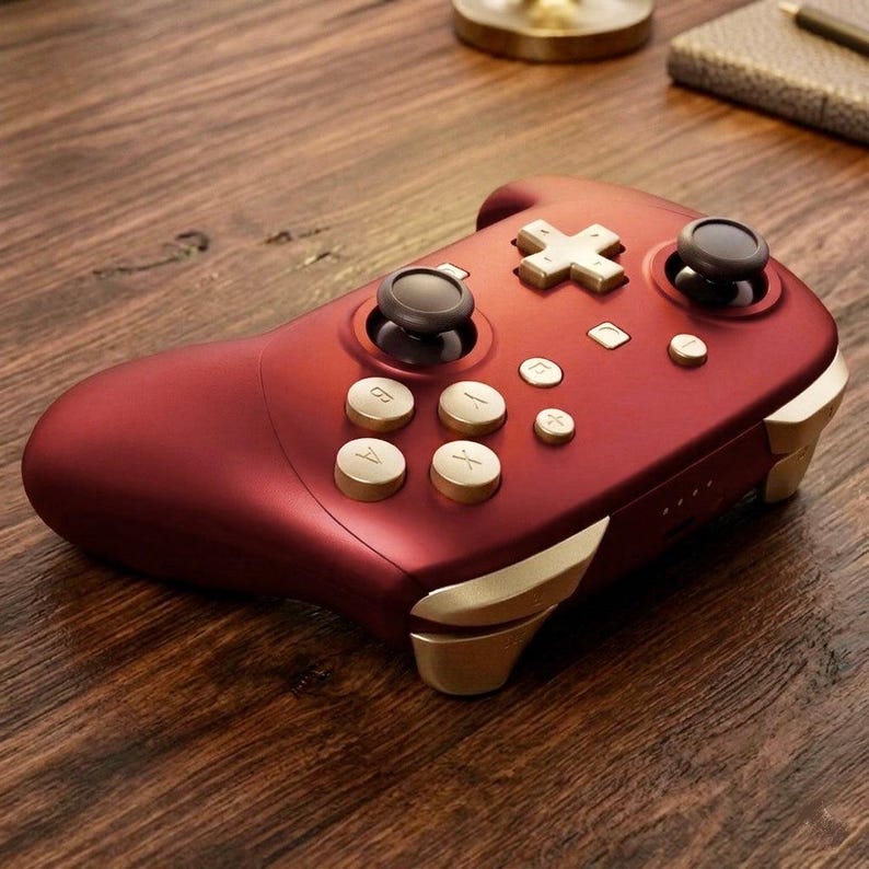 May include: A matte dark red wireless video game controller featuring gold-colored buttons, a gold directional pad, and two black analog sticks. The controller includes labeled A, B, X, and Y buttons, along with gold-toned shoulder triggers.