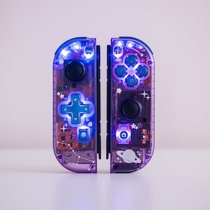 May include: A pair of purple and blue Nintendo Switch Joy-Con controllers with a galaxy theme. The controllers have a clear plastic shell with stars, planets, and constellations printed on them. The buttons are teal and the lights are purple.
