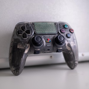 May include: A transparent gray video game controller with black buttons and joysticks. The controller has colored action buttons and a PlayStation logo. The design allows a view of the internal components.