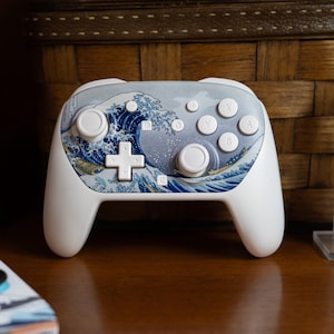 May include: White video game controller featuring a blue and white wave design, reminiscent of the famous Japanese artwork. The controller has a cross-shaped directional pad, four face buttons, and two analog sticks. The buttons are labeled with letters.