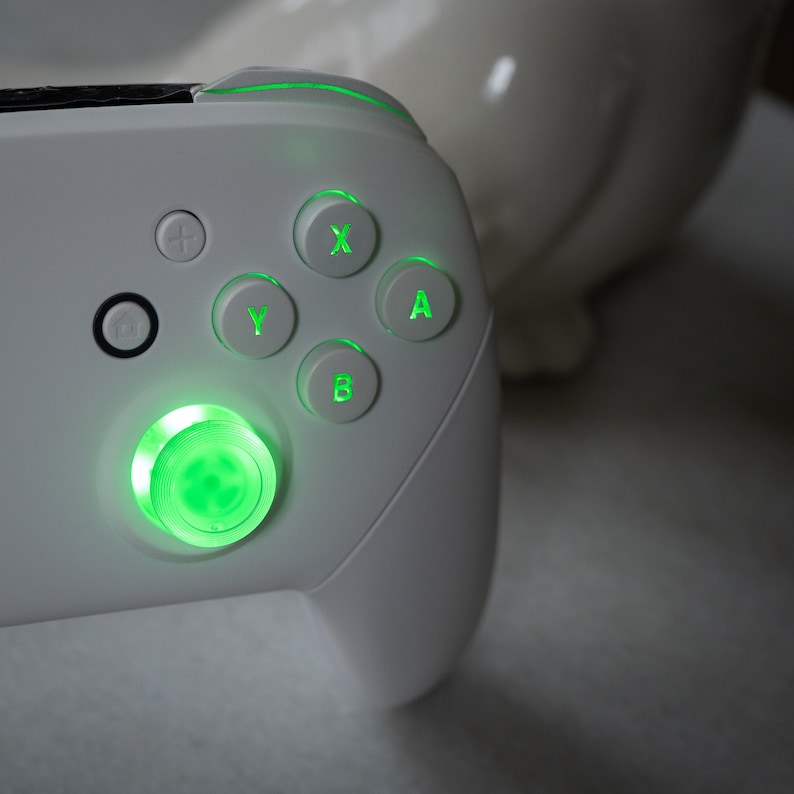May include: A white gaming controller with green illuminated buttons. The controller has a circular button with a green light in the center, and the buttons labeled A, B, X, and Y are also illuminated in green.