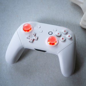 May include: A white Nintendo Switch controller with red buttons. The controller has a D-pad, four face buttons labeled X, Y, B, and A, and two analog sticks.
