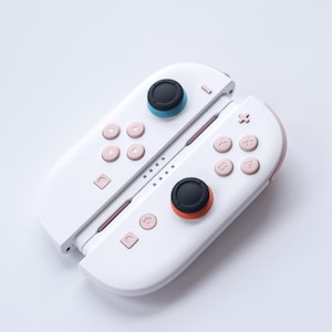 May include: White and pink Nintendo Switch Joy-Con controllers. The controllers have pink buttons and accents, with black and teal or orange joysticks. The controllers are shown side by side.