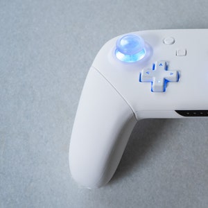 May include: A white video game controller with a blue light illuminating the buttons and the analog stick.