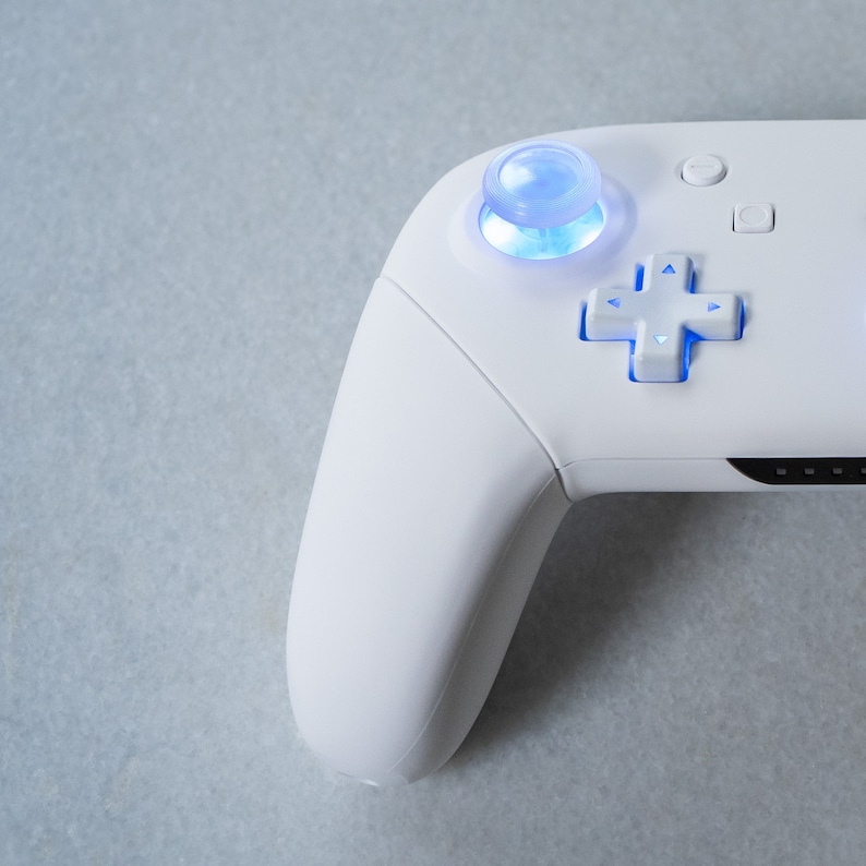 May include: A white video game controller with a blue light illuminating the buttons and the analog stick.
