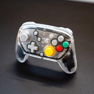May include: A transparent video game controller with a variety of colored buttons, including yellow, red, green, and white. The controller has a directional pad and analog stick. The design is modern and sleek, suitable for gaming enthusiasts.
