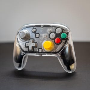May include: A transparent video game controller with a clear plastic shell. It features gray, yellow, red, and green buttons, a gray directional pad, and gray analog sticks. The controller is designed for gaming.