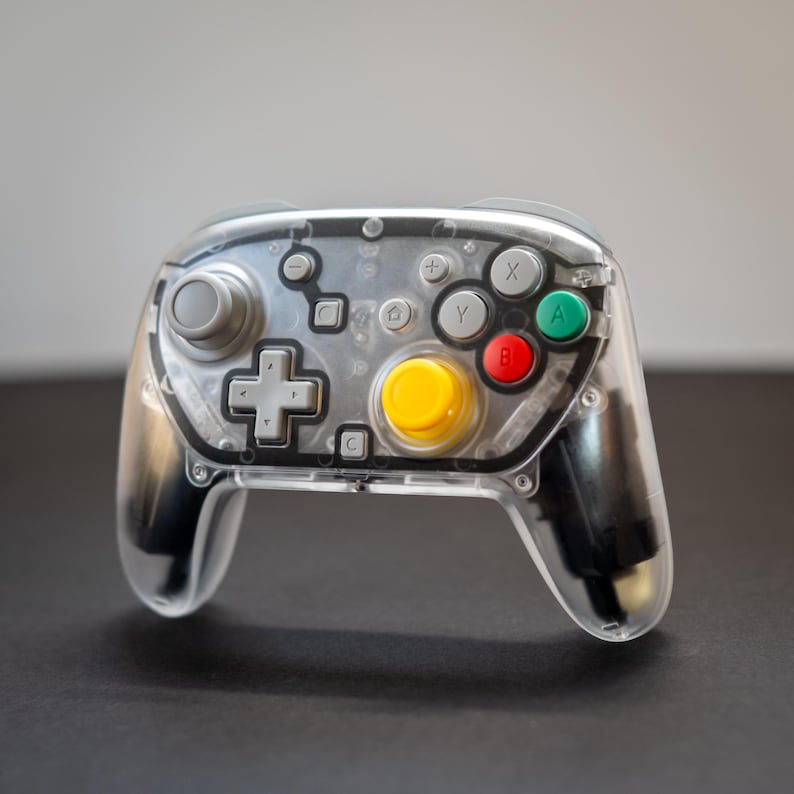 May include: A transparent video game controller with a clear plastic shell. It features gray, yellow, red, and green buttons, a gray directional pad, and gray analog sticks. The controller is designed for gaming.