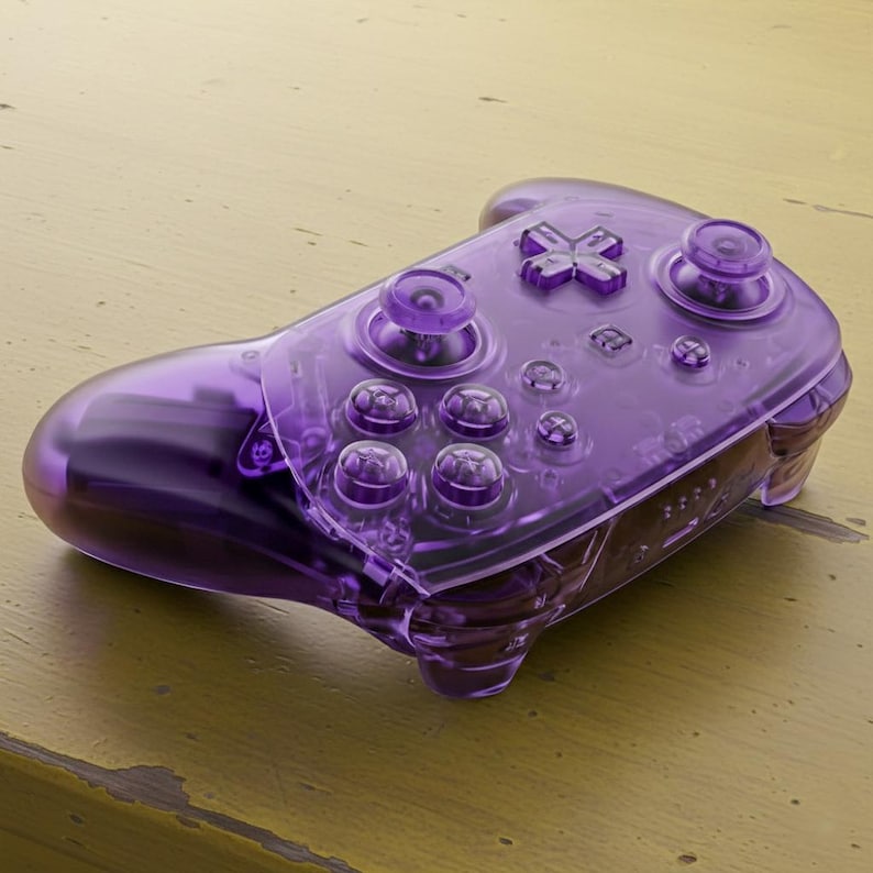 Puede incluir: Un mando de videojuegos morado transparente con botones, cruceta y joysticks. El mando est&aacute; hecho de un material transparente y brillante, y est&aacute; dise&ntilde;ado para jugar.
