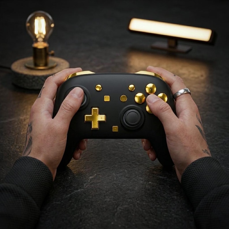 May include: Black and gold gaming controller with a cross-shaped directional pad and multiple gold buttons. The controller is held in both hands, with a ring on one finger. The background includes a lamp and a light bar.