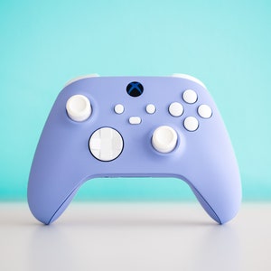Puede incluir: Un mando Xbox de color morado lavanda con botones blancos y un pad direccional blanco. El mando tiene un logotipo de Xbox azul en la parte delantera.