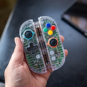 May include: A clear, transparent video game controller with visible internal circuitry. The controller features colored buttons, a directional pad, and analog sticks. The design is modern and sleek, with a focus on the internal components. The controller is held in a hand.