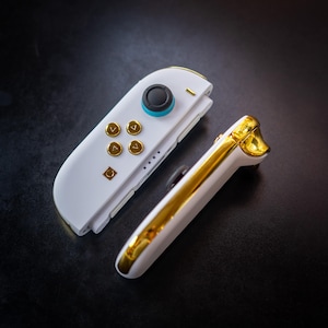 以下が含まれることがあります： 白と金のビデオゲームコントローラー。コントローラーは、金色のアクセントとボタンが付いた白いベースです。ジョイスティックは、青いリングが付いた黒いトップです。コントローラーは暗い表面にあります。
