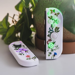 May include: Two white Nintendo Switch Joy-Con controllers with floral designs. The left controller has a purple button and the right controller has green buttons. The controllers are on a white surface.