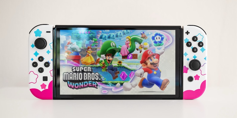 Pode incluir: Um console Nintendo Switch branco com Joy-Cons rosa e azul com um padr&atilde;o de estrelas e nuvens. O console est&aacute; exibindo o jogo Super Mario Bros. Wonder.