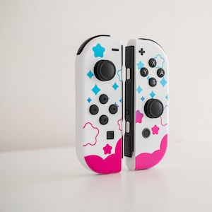 Pode incluir: Um controlador Nintendo Switch Joy-Con branco e rosa com um design colorido com estrelas e nuvens. O controlador tem um bot&atilde;o preto e um joystick preto.