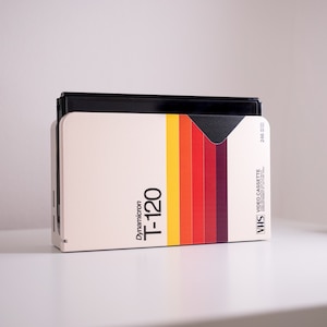 Puede incluir: Una cinta de video VHS vintage con un diseño de rayas de arcoíris. La cinta está etiquetada como "VHS VIDEO CASSETTE" y "Dynamicron T-120".
