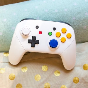 May include: White wireless video game controller with black accents and colorful buttons. The controller has a blue, yellow, and red button on the top, and a white button on the front.