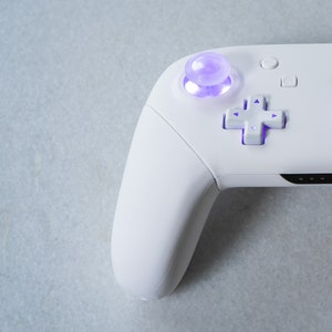 May include: A white video game controller with a purple button and a white directional pad. The controller is on a light gray surface.
