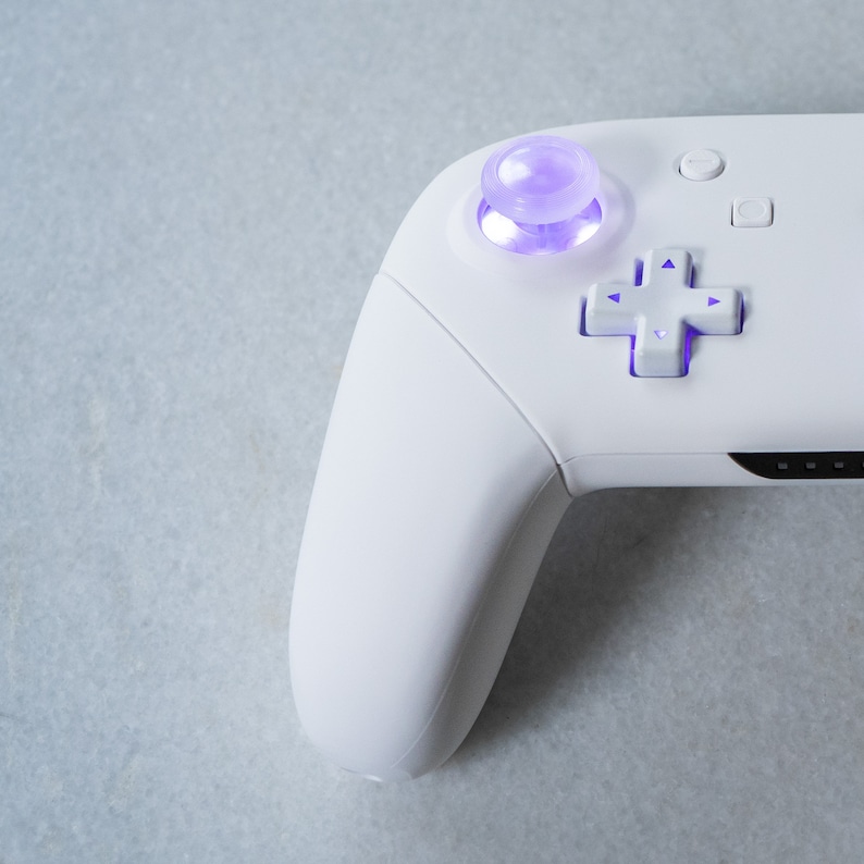 May include: A white video game controller with a purple button and a white directional pad. The controller is on a light gray surface.