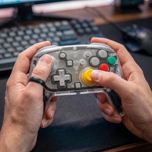May include: A clear, transparent video game controller with gray buttons and a yellow joystick. The controller has a plus-shaped directional pad and colored buttons, including red, green, and yellow. The controller is held in two hands.