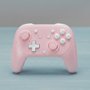 Puede incluir: Un mando de videojuegos inalámbrico rosa con botones y detalles en blanco. El mando cuenta con dos palancas analógicas, una cruceta y varios botones de acción. El diseño es simétrico, con una forma suave y ergonómica.