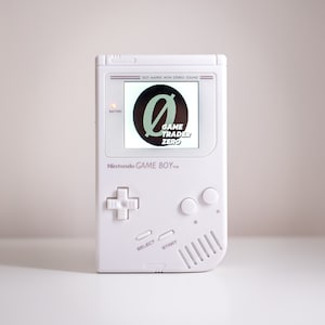 Puede incluir: Una consola de juegos portátil Nintendo Game Boy blanca con una pantalla en blanco y negro que muestra el texto "GAME TRADER ZERO" y un gran símbolo de cero. La consola tiene una luz de encendido roja, un pad direccional blanco y botones blancos etiquetados como "A", "B", "START" y "SELECT".