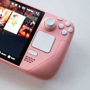 Könnte beinhalten: Eine rosa Handheld-Spielkonsole mit einem Bildschirm, auf dem Spieletitel angezeigt werden. Die Konsole verfügt über weiße Tasten, einen weißen Joystick und ein weißes Touchpad. Der Bildschirm zeigt Spiele wie "Hollow Knight" und "Bloodline".