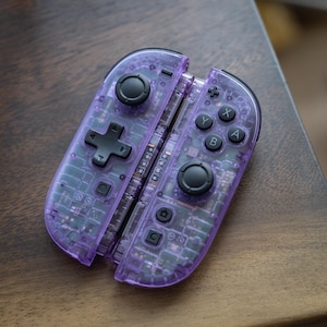 Op de afbeelding: Een paar doorzichtige paarse videogamecontrollers met een zichtbare interne printplaat, zwarte analoge sticks, een richtingspad en standaard actieknoppen met de labels A, B, X en Y, samen met home- en capture-functieknoppen.