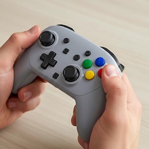 Könnte beinhalten: Ein grauer Videospiel-Controller mit schwarzen Joysticks, einem Steuerkreuz und Tasten. Der Controller hat farbige Knöpfe in Grün, Blau, Rot und Gelb. Der Controller wird mit zwei Händen gehalten.