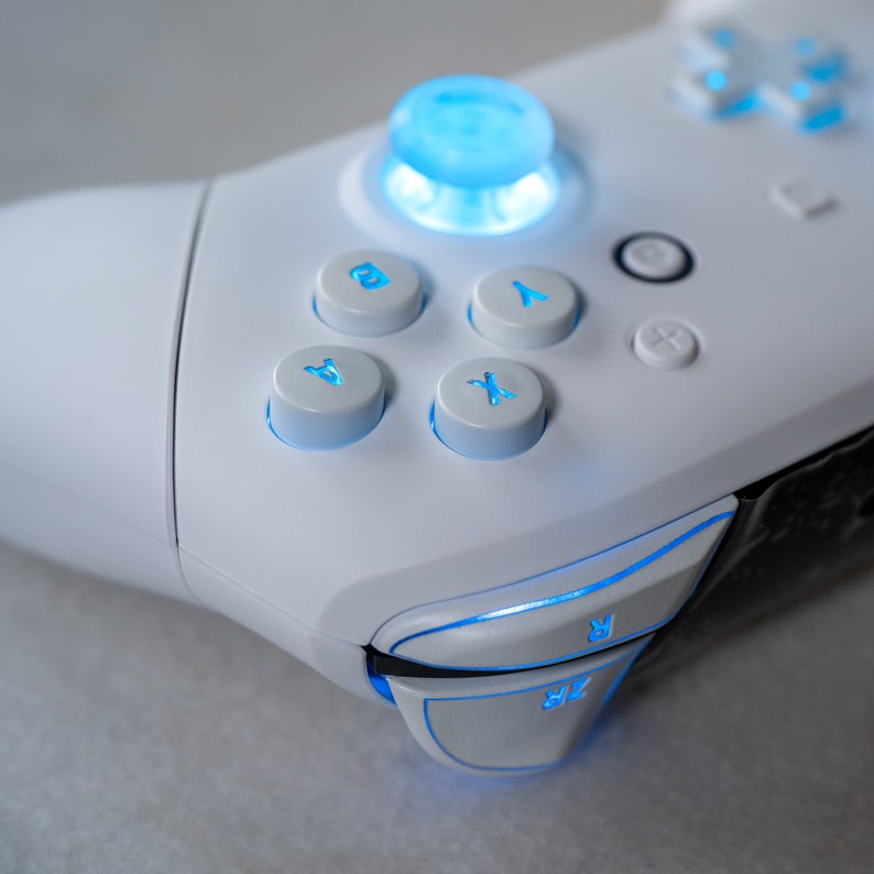 May include: A white gaming controller with blue illuminated buttons. The controller has a circular button in the center, four face buttons labeled A, B, X, and Y, and two shoulder buttons labeled R and ZR.