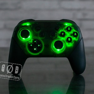 May include: A black Nintendo Switch controller with green LED lights around the buttons and joysticks. The controller is on a reflective surface.