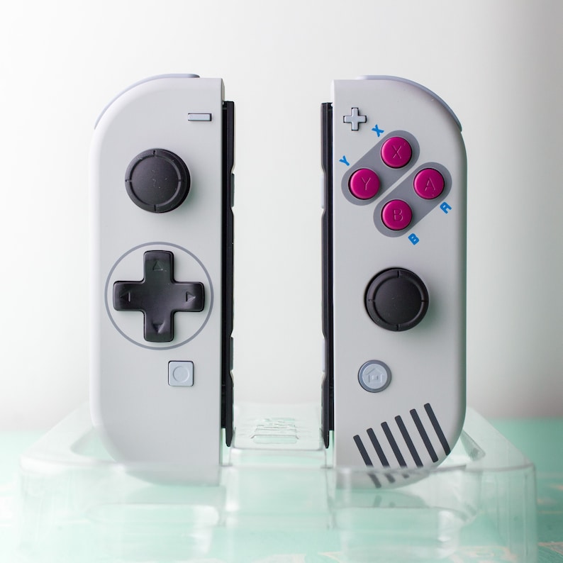 Może przedstawiać: Para białych kontroler&oacute;w Joy-Con do Nintendo Switch z czarnymi przyciskami i szaro-białym paskowanym wzorem na prawym kontrolerze.