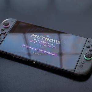 Puede incluir: Consola Nintendo Switch con el juego "Metroid Prime 4 Beyond" en la pantalla. Los mandos negros tienen detalles rosas y verdes. La pantalla muestra el t&iacute;tulo del juego y "Nintendo Switch 2 Edition".