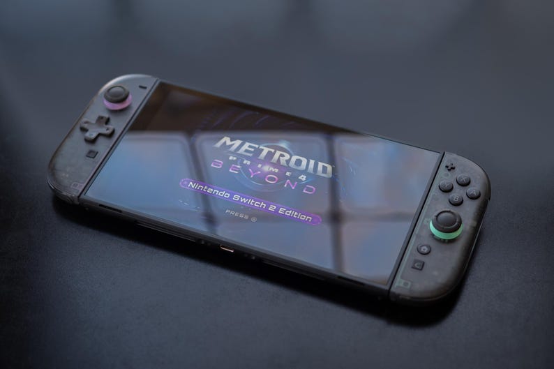 Puede incluir: Consola Nintendo Switch con el juego "Metroid Prime 4 Beyond" en la pantalla. Los mandos negros tienen detalles rosas y verdes. La pantalla muestra el t&iacute;tulo del juego y "Nintendo Switch 2 Edition".