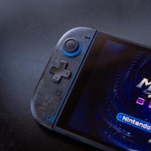 Puede incluir: Una consola de juegos port&aacute;til con una carcasa de mando gris oscuro transl&uacute;cida que deja ver los circuitos internos. El dispositivo incluye una cruceta negra, un joystick circular con una base azul brillante y una pantalla con gr&aacute;ficos.