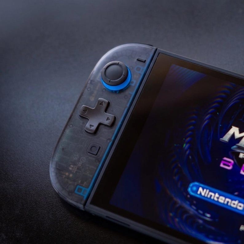 Puede incluir: Una consola de juegos port&aacute;til con una carcasa de mando gris oscuro transl&uacute;cida que deja ver los circuitos internos. El dispositivo incluye una cruceta negra, un joystick circular con una base azul brillante y una pantalla con gr&aacute;ficos.