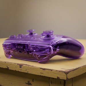 Puede incluir: Un mando de videojuegos p&uacute;rpura transparente con componentes internos visibles. El mando tiene dos palancas anal&oacute;gicas, una cruceta y varios botones. El dise&ntilde;o es elegante y moderno, con un acabado brillante. El mando est&aacute; sobre una superficie de madera.