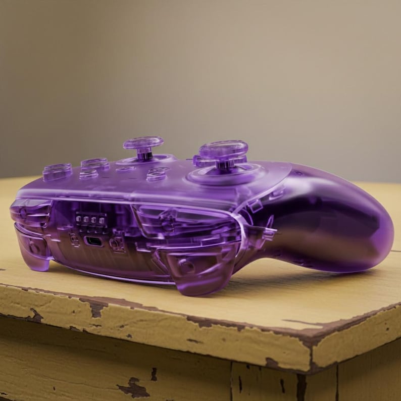 Puede incluir: Un mando de videojuegos p&uacute;rpura transparente con componentes internos visibles. El mando tiene dos palancas anal&oacute;gicas, una cruceta y varios botones. El dise&ntilde;o es elegante y moderno, con un acabado brillante. El mando est&aacute; sobre una superficie de madera.
