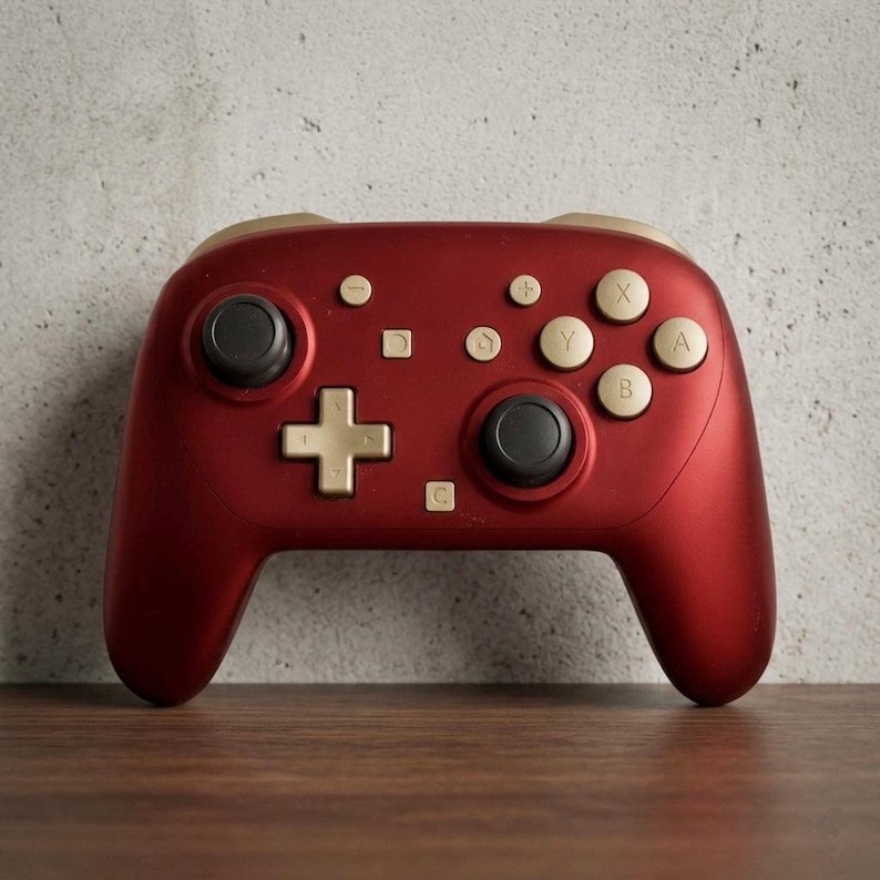 May include: A matte dark red wireless video game controller featuring gold-colored buttons, a gold directional pad, and two black analog sticks. The controller includes labeled buttons with the letters A, B, X, and Y, along with symbols for plus, minus, and home.