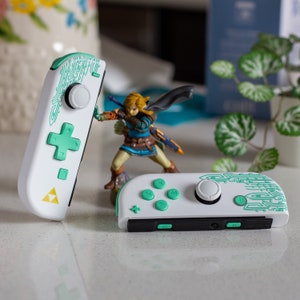 Pode incluir: Dois controladores brancos do Nintendo Switch com detalhes verdes e azuis. Um controlador tem um design verde e azul com um botão circular e um direcional. O outro controlador tem um design verde e azul com um botão circular e quatro botões.